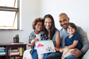multi-cultural-family-sitting-on-couch-smiling-2-parents-2-kids-holding-drawing-of-a-house